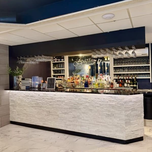 A modern bar with a white stone facade, black countertop, and shelves stocked with bottles and glasses.