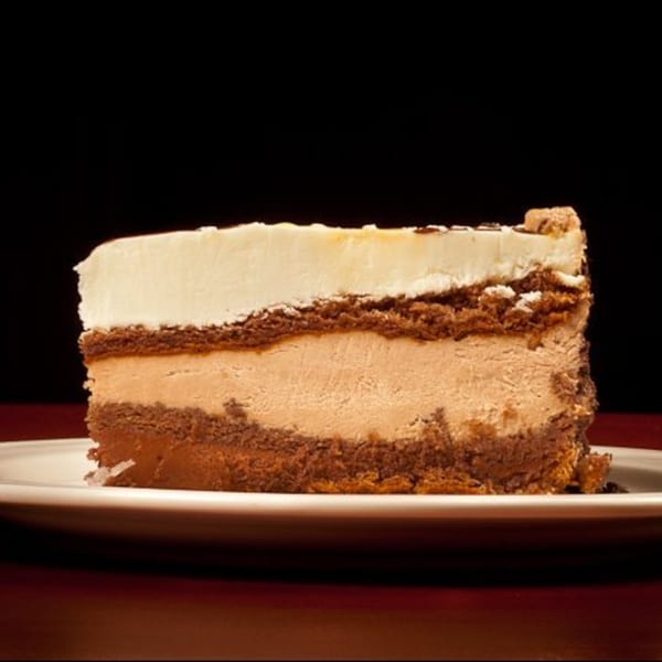 A slice of layered chocolate and caramel cheesecake with a creamy white topping on a white plate.