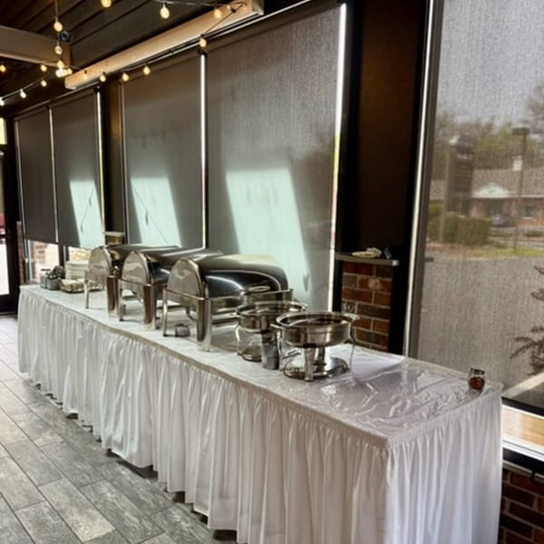 A buffet table with chafing dishes and serving utensils, set up in a room with large windows.