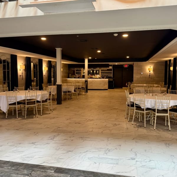 An empty restaurant dining room with round tables, white tablecloths, and a bar in the background.