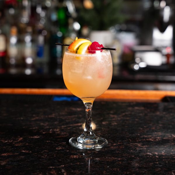 A light orange cocktail in a stemmed glass, garnished with orange slices and cherries, on a dark bar.