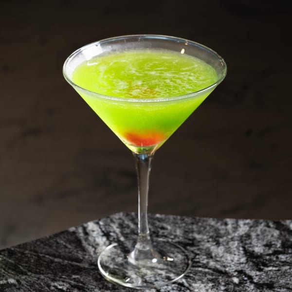 Bright green cocktail with a red cherry at the bottom, served in a martini glass on a dark marble surface.