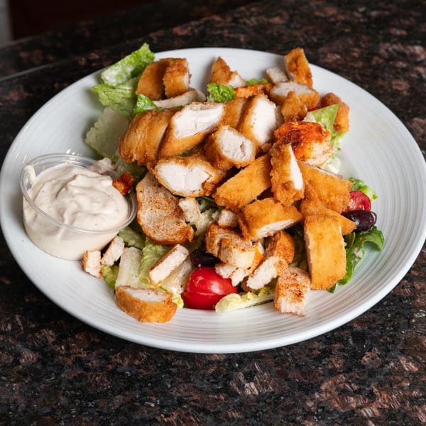 Crispy chicken salad with lettuce, tomatoes, olives, croutons, and a side of dressing.