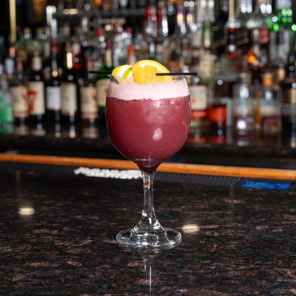 A purple cocktail with white foam and orange slices sits on a dark bar counter with liquor bottles in the background.