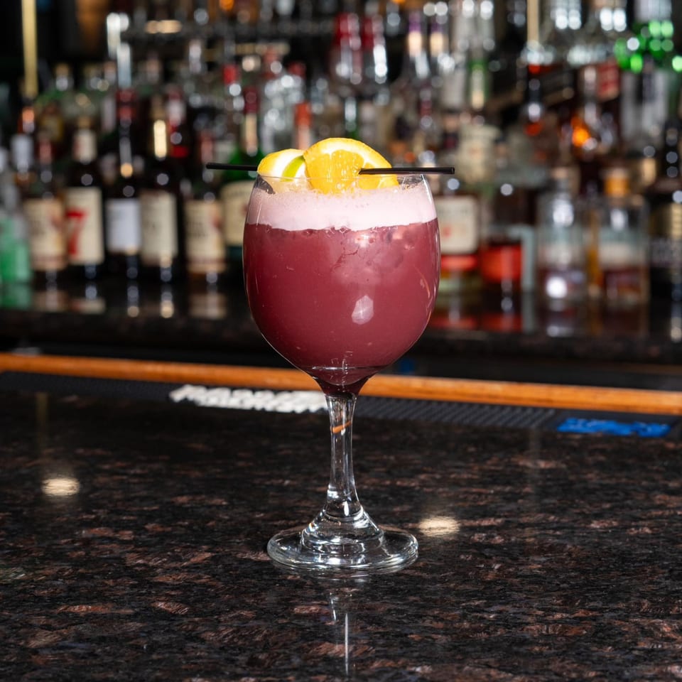 A purple cocktail with white foam and orange slices sits on a dark bar counter with liquor bottles in the background.