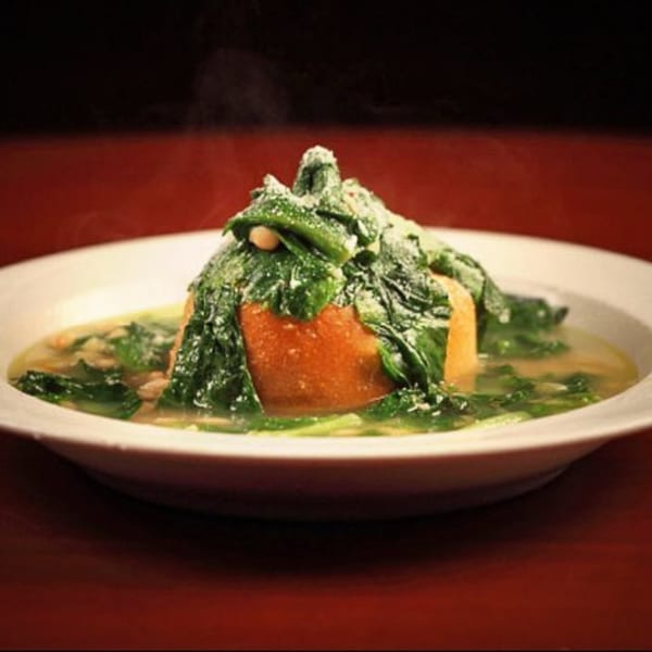 Steaming hot bowl of soup with a large piece of bread covered in green vegetables and cheese.