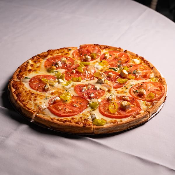 A whole pizza with sliced tomatoes, green peppers, and feta cheese on a white tablecloth.