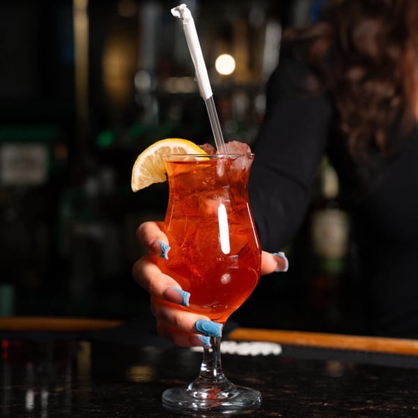 A bartender's hand with blue nails holding a red cocktail with a lemon slice and straw.